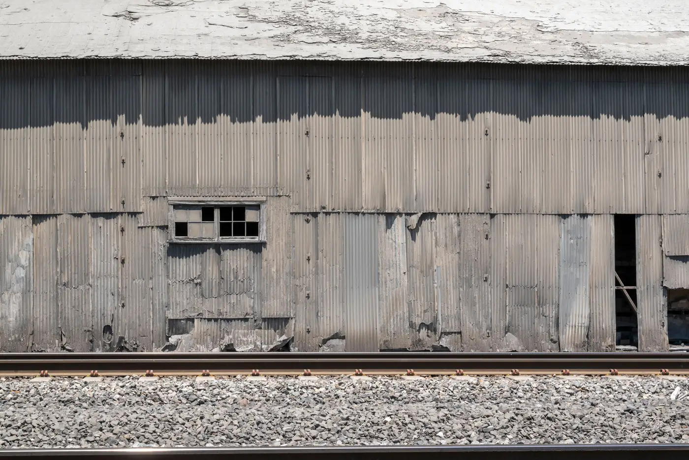 Weathered corrugated-metal industrial building wall beside a rail spur with gravel ballast track in the foreground.